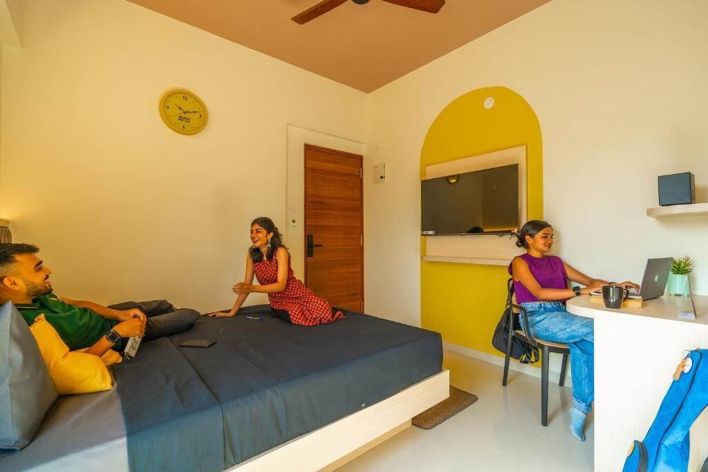 A room with a bed, a desk, and a chair. There is a clock on the wall and a window with a view.