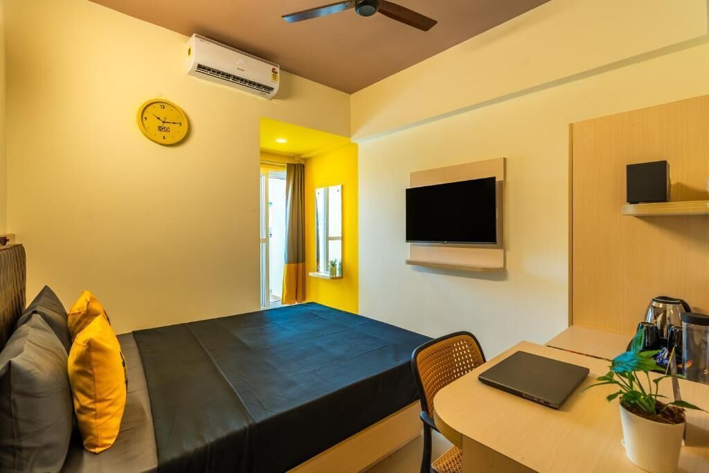 Una habitación de hotel compacta con una cama, un escritorio, una silla, un televisor y un equipo de aire acondicionado.