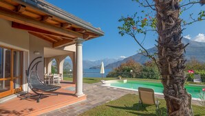 A poolside area with a lounge chair, a table, and a view of a mountain lake. - My Rental Homes - Villa Cremia  with infrared sauna, Jacuzzi and lake view (Vignola)