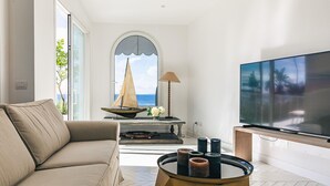 A modern living room with a sofa, a television, a table with candles, a lamp, and a view of the sea through a window. - Villa Heart with private pool - My Rental Homes (Massa Lubrense)