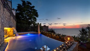 A pool with a waterfall feature, surrounded by stone walls and plants, overlooking a sunset view of the sea. - Villa Heart with private pool - My Rental Homes (Massa Lubrense)