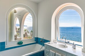 Ett badrum med ett stort badkar, ett handfat och två välvda fönster med utsikt över havet.