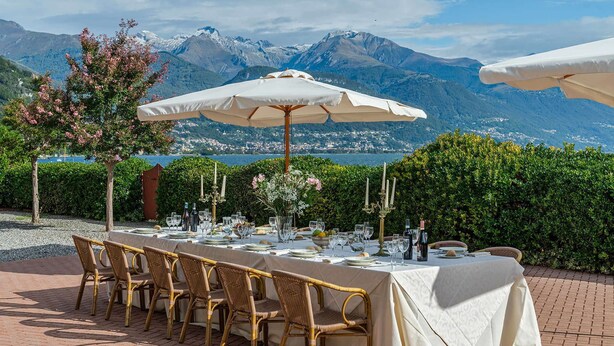 Mobiliario de comedor exterior con una mesa dispuesta para seis personas, rodeado de sillas de mimbre, y una vista a montañas y una masa de agua al fondo.