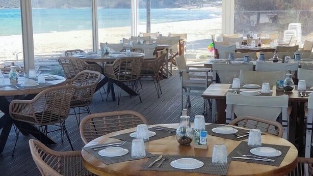 Een buitenruimte met een eethoek, rieten stoelen en tafels, en grote ramen met uitzicht op het strand en de zee.
