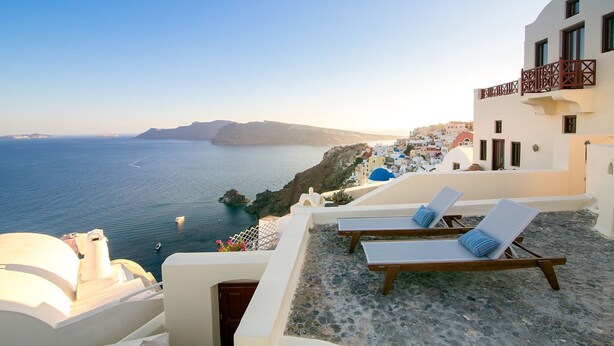 A seaside hotel with white buildings, blue-domed roofs, and a view of the sea. There are lounge chairs with blue cushions on a terrace. - My Rental Homes - Palea Suite with Jacuzzi, sea view e wonderful rooftop terrace (Fira)