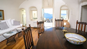 A dining area with a wooden table, chairs, and a decorative bowl. There is a sofa with cushions and a small table with a book. The room has white walls and a view of the sea through an open door.