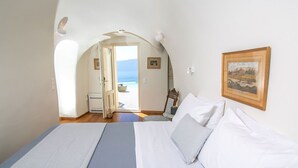 A bedroom with a bed, a chair, a framed picture, and a view of the sea through an open door.