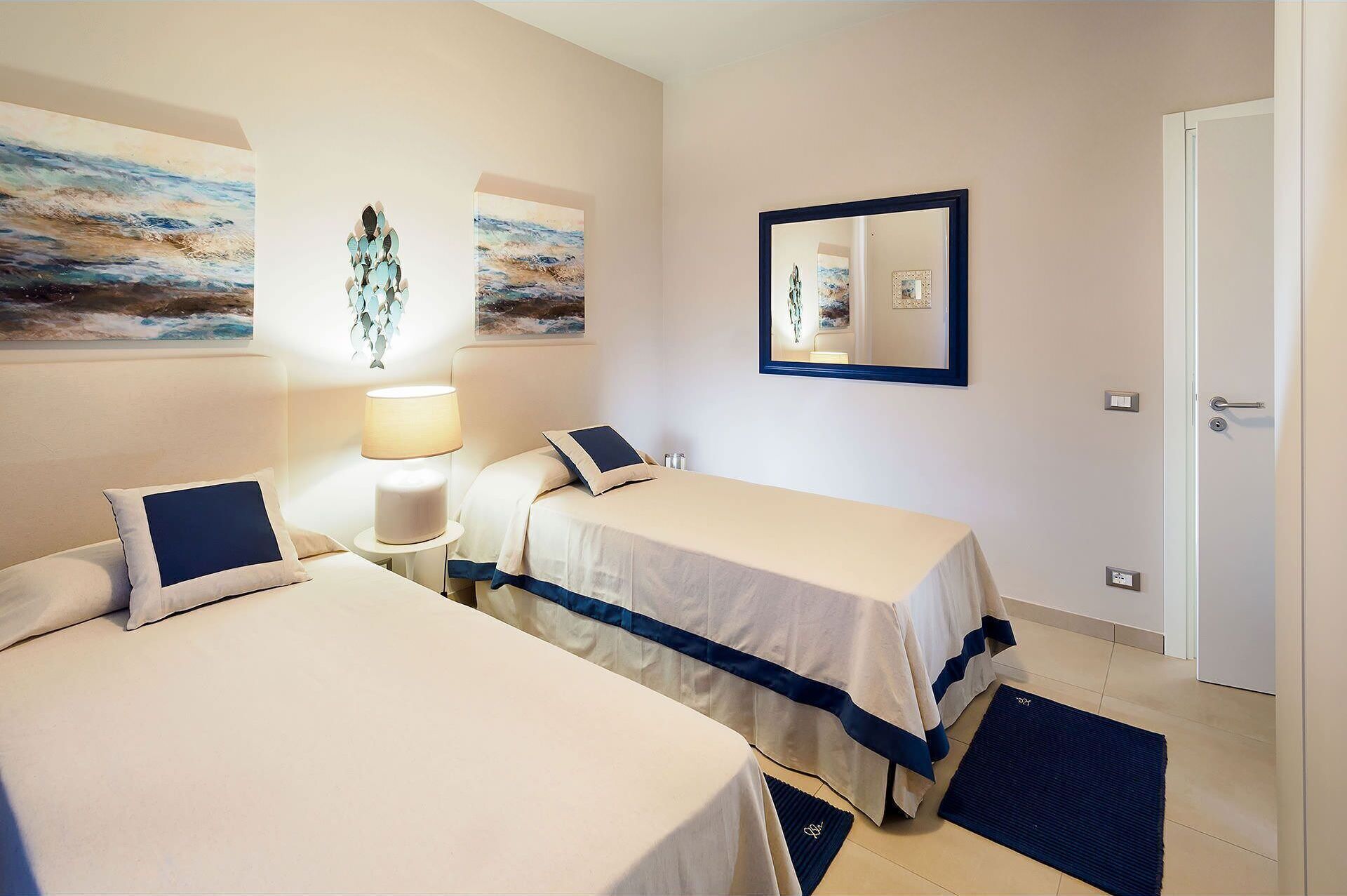 Une chambre d’hôtel avec deux lits, une table de chevet avec une lampe, des œuvres d’art aux murs et une porte.