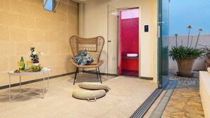 A corner of a hotel room with a wicker chair, a small table with drinks and flowers, and a red shower area visible through a glass door. - Sun Stone with garden and pool - My Rental Homes (Trapani)