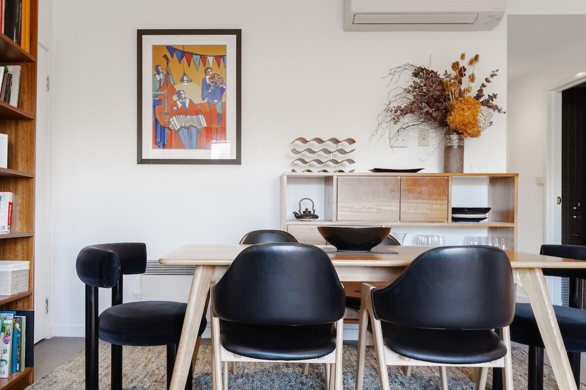 Un espace repas moderne avec une table en bois, des chaises noires, une œuvre d’art encadrée et un vase décoratif contenant des fleurs séchées.
