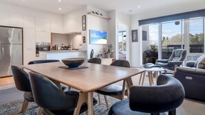 Une cuisine moderne avec des meubles blancs, un coin repas doté d’une table en bois et de chaises noires, et un salon avec un canapé et des fauteuils.