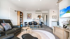 Un salon moderne avec un canapé en cuir noir, un coin repas avec une table et des chaises en bois, et un grand tableau représentant une scène de plage accroché au mur.