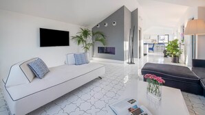 Een moderne woonkamer met een witte bank, een zwarte bank en een salontafel met tijdschriften. Er staan twee potplanten en een tv is aan de muur bevestigd.
