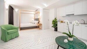 A modern hotel room with a bed, a green armchair, a small dining table, and a kitchen area with white cabinets and a sink.