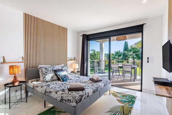 A modern bedroom with a bed, a nightstand, a lamp, a TV, and a balcony with a table and chairs. - Chambre "La Garden" - Heated pool & SPA (Plaissan)