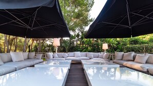 A modern outdoor seating area with a glass-top table and cushioned sofas, surrounded by umbrellas and lush greenery. - My Rental Homes - Villa Sound luxury property with pool and privacy (San Jose)