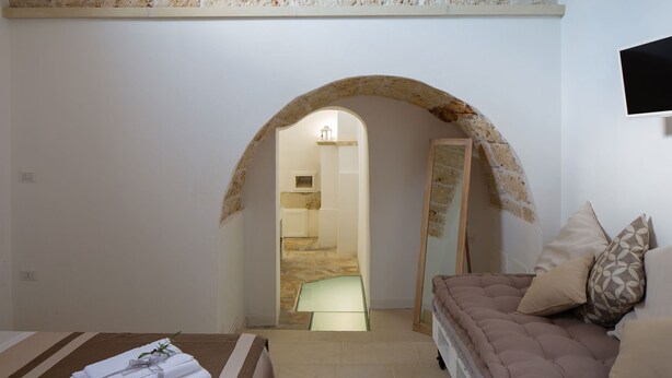 A room with a stone archway leading to a staircase, a bed with a brown cover, and a TV mounted on the wall. - My Rental Homes - Villa Salentina with private pool and large private garden (Cutrofiano)