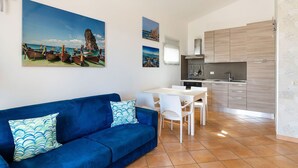 A modern kitchen with wooden cabinets, a stove, and a sink. There is a blue sofa with patterned pillows and a dining table with white chairs. - Apartment Ippocampo with terrace - My Rental Homes (Noto)