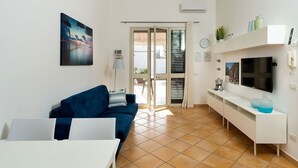 Una sala moderna con un sofá azul, una mesa de centro blanca, un televisor y una zona de cocina con un armario blanco y un jarrón azul.
