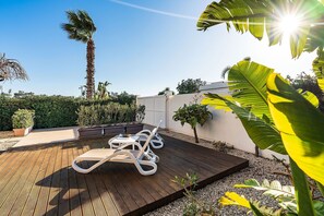 A wooden deck with lounge chairs, surrounded by tropical plants and a palm tree. - Apartment Kalimba with terrace - My Rental Homes (Noto)