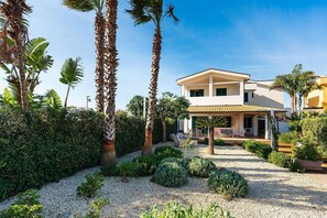 A two-story house with a covered patio, surrounded by palm trees and a gravel pathway.