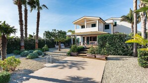 A two-story house with a covered patio, surrounded by palm trees and a gravel pathway. - Stella Marina near the sea  - My Rental Homes (Noto)