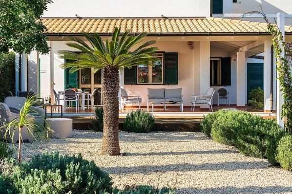 A covered outdoor seating area with a palm tree, white furniture, and a gravel ground.