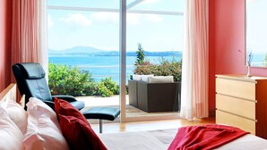 A bedroom with a bed, a chair, a dresser, and a view of the sea.
