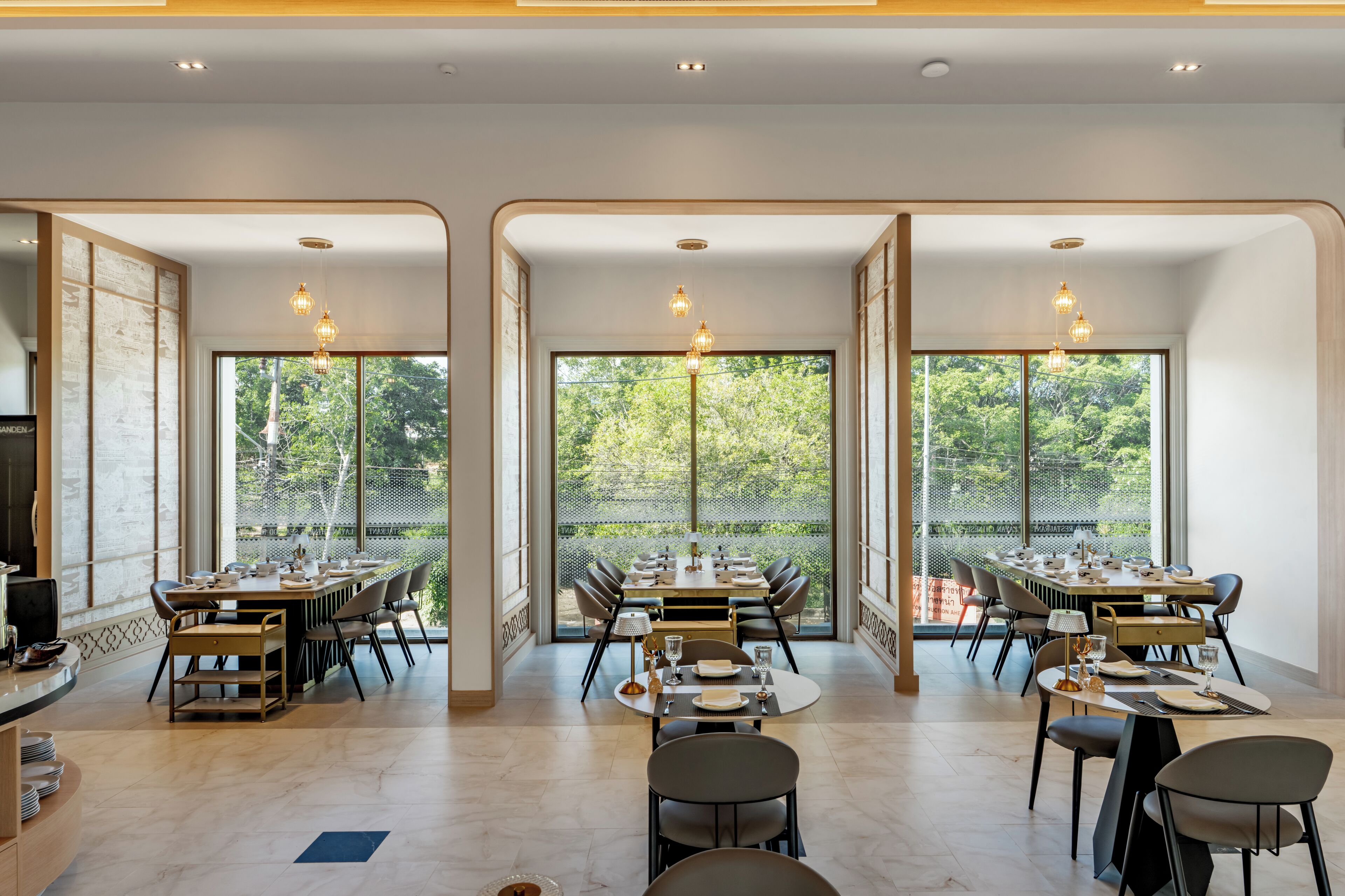 A modern dining area with multiple tables and chairs, large windows with a view of greenery, and pendant lighting.