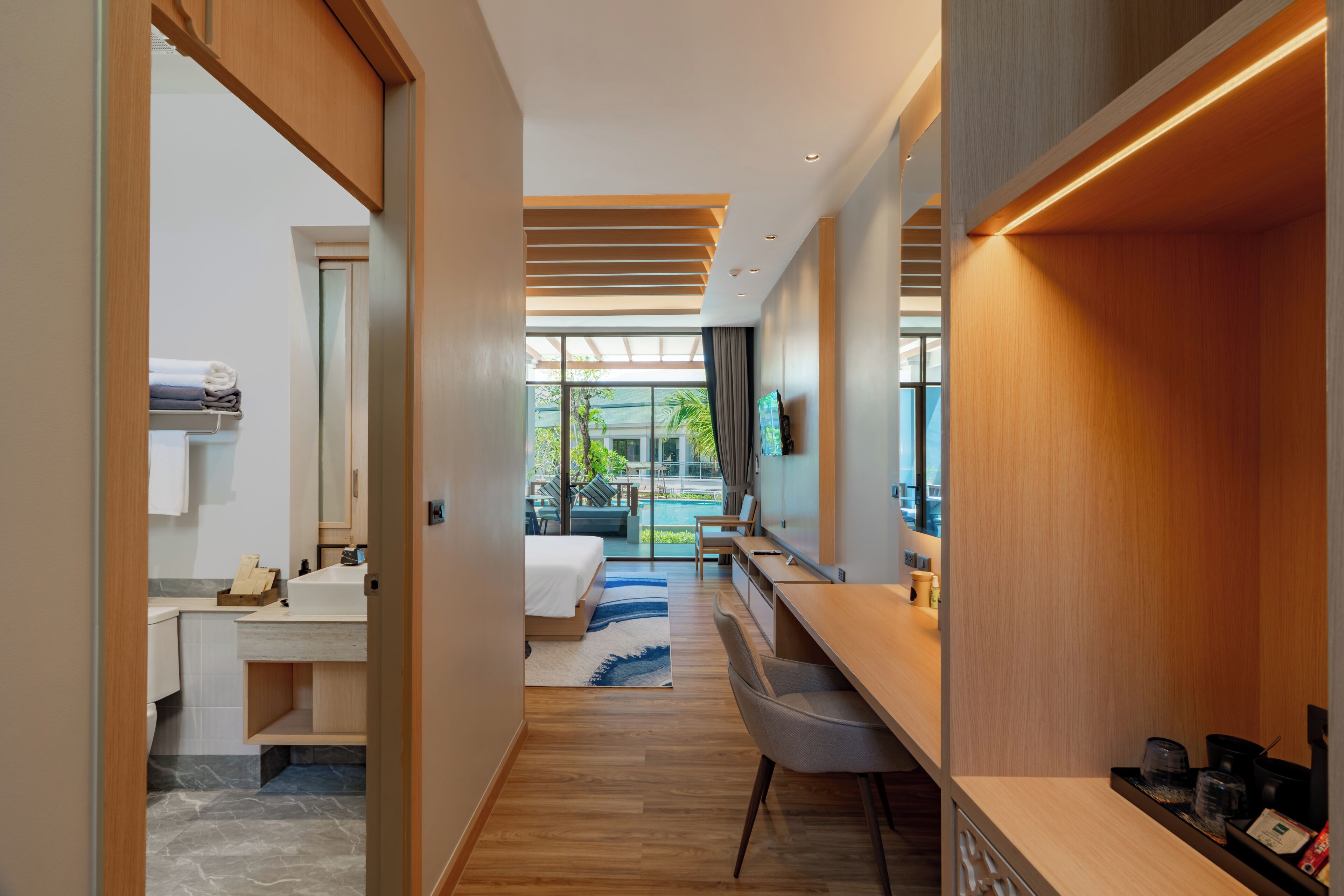 Ein modernes Hotelzimmer mit Holzboden, einem Bett mit weißer Bettdecke, einem Schreibtisch mit Stuhl, einem Badezimmer mit Waschbecken und Spiegel sowie Blick auf einen Pool und Grünflächen.
