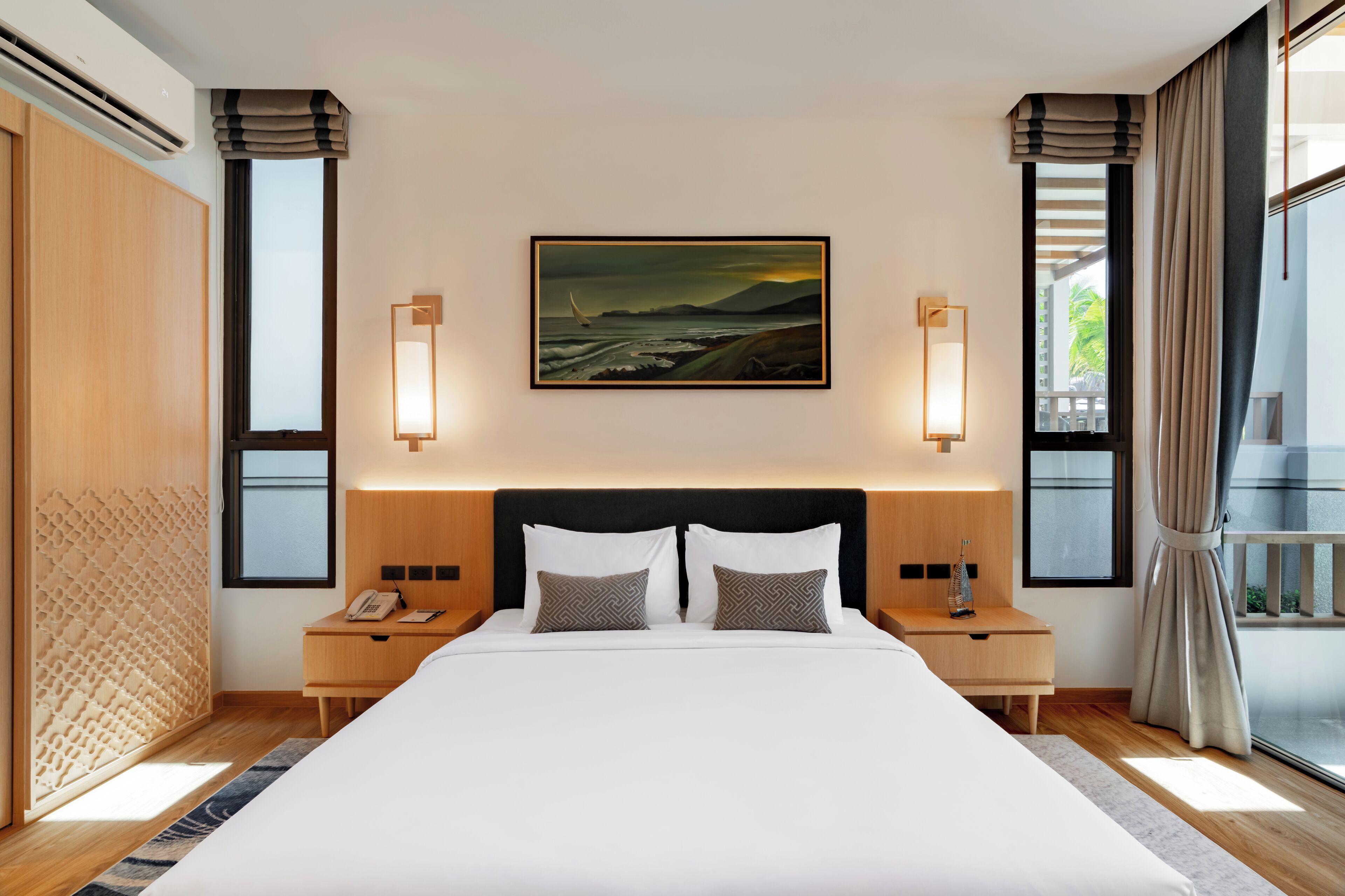 A hotel room with a large bed, two bedside tables, a painting above the bed, and a view of the outdoors through the windows.