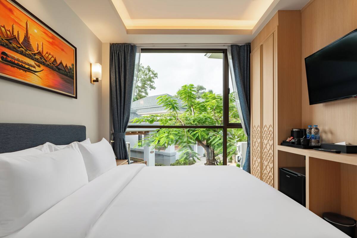 A hotel room with a large bed, a TV mounted on the wall, a window with a view of greenery, and a painting on the wall.