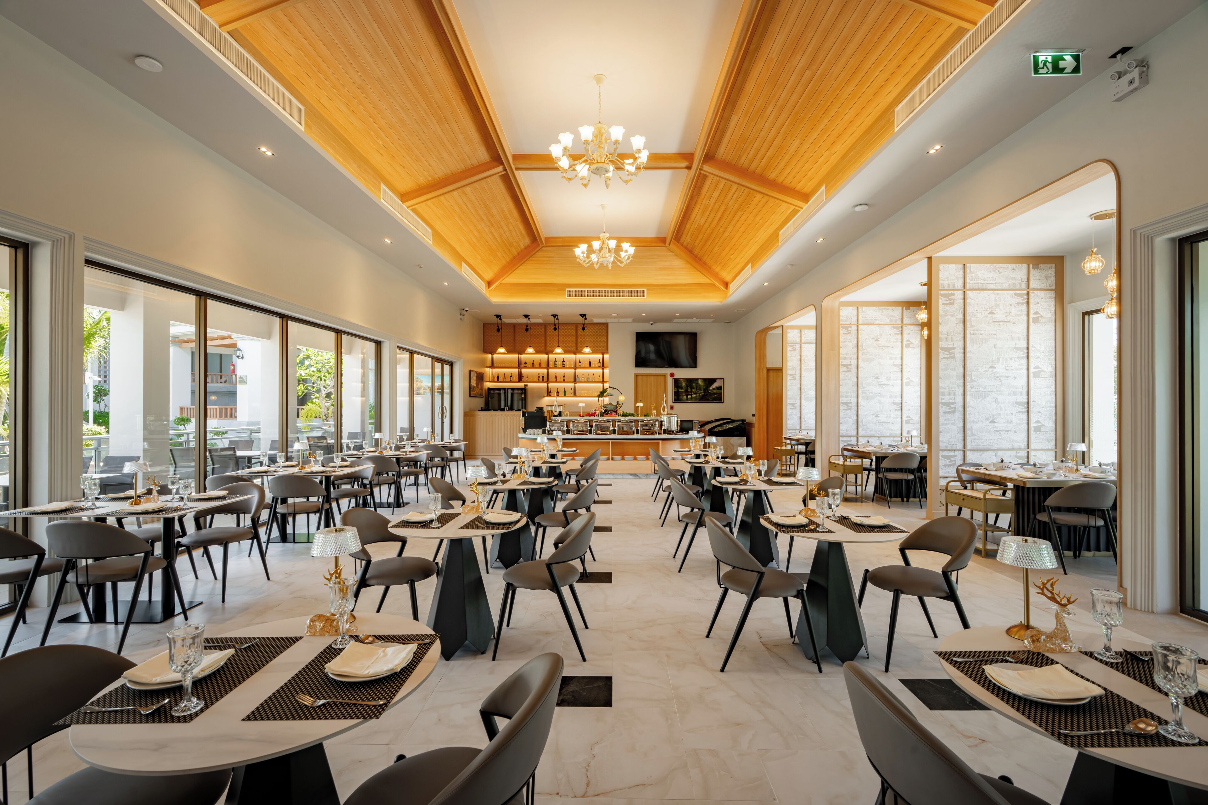 A spacious dining area with multiple tables and chairs, a central bar area, and large windows providing natural light.