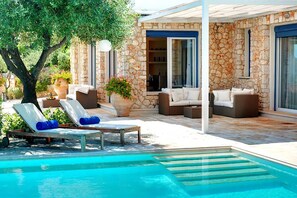 A poolside area with lounge chairs and a stone building with large windows. - Villa Azzurra with private pool - My Rental Homes (Corfu)