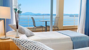 Une chambre d’hôtel avec deux lits, une table de chevet, une lampe, un vase décoratif contenant des branches et une vue sur la mer par de grandes fenêtres.