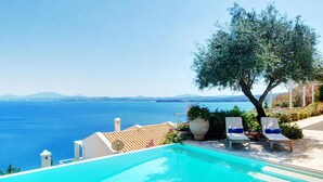 Une piscine offrant une vue dégagée sur la mer, deux chaises longues et un grand arbre.