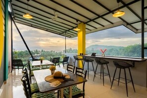 A dining area with a table and chairs, a bar area with stools, and a view of the landscape. - The Hosteller Gokarna (Kumta)