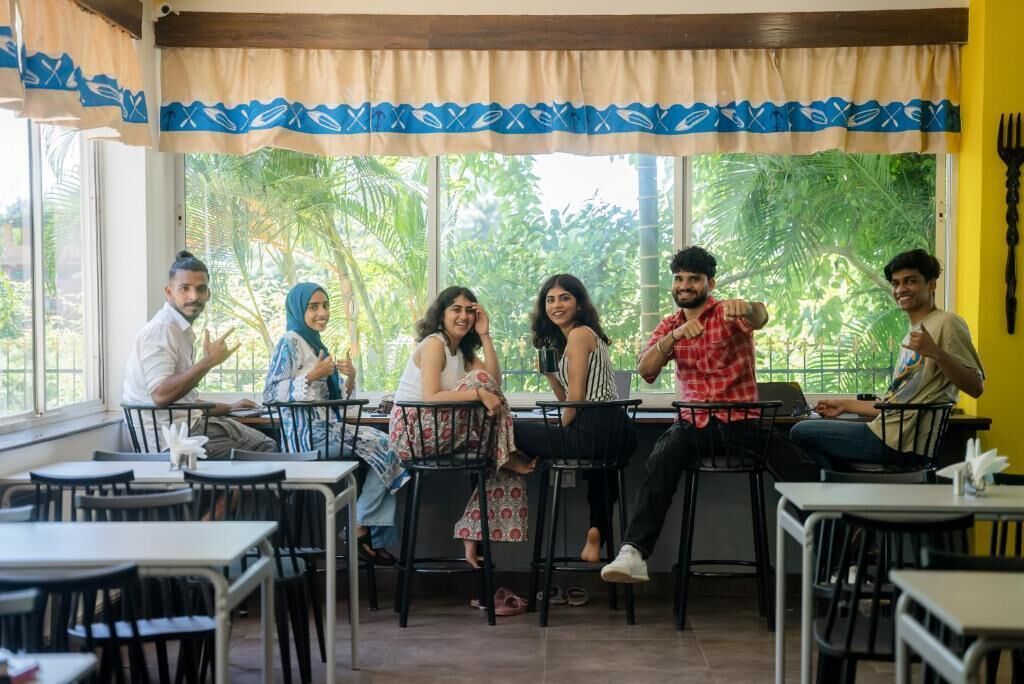 A group of people seated at tables in a cafe with large windows and a view of greenery outside.