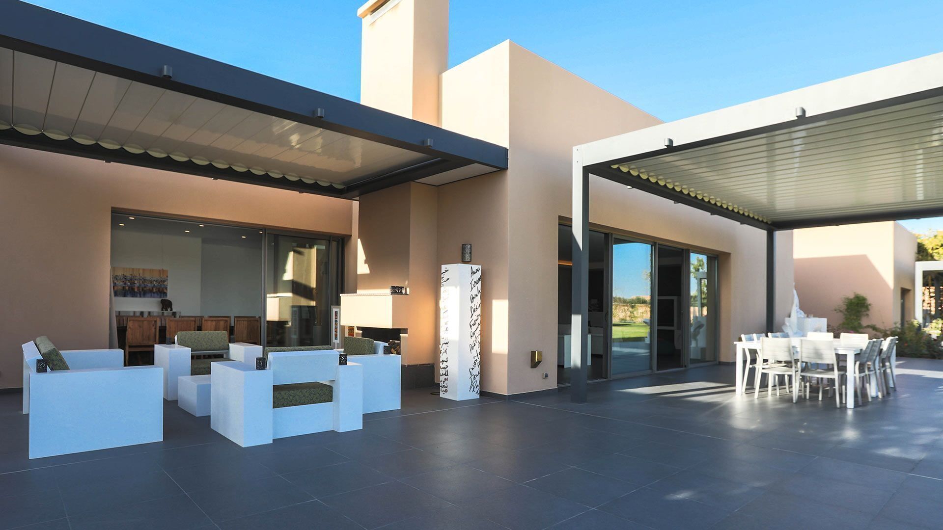 A modern outdoor patio with white furniture, a covered seating area, and a large sliding glass door leading to a garden.