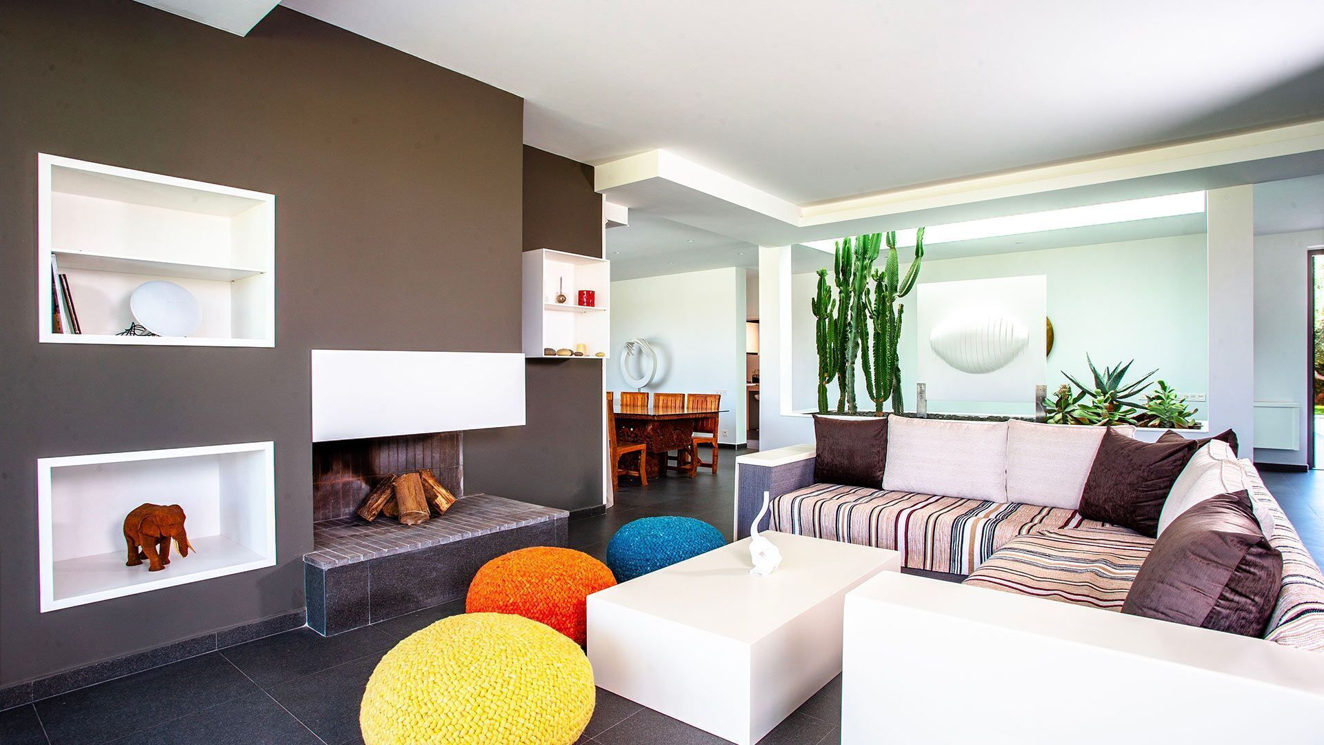 Ein modernes Wohnzimmer mit Kamin, bunten Poufs, einem gestreiften Sofa und einem Essbereich im Hintergrund.
