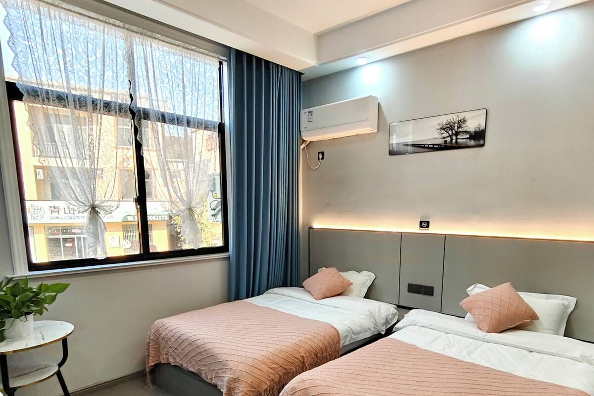 Habitación de hotel con dos camas, una mesita, una planta en maceta y un ventanal con cortinas.