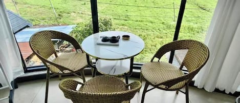A room with a large window, wicker chairs, a small table with a tray, and a view of green fields and mountains.