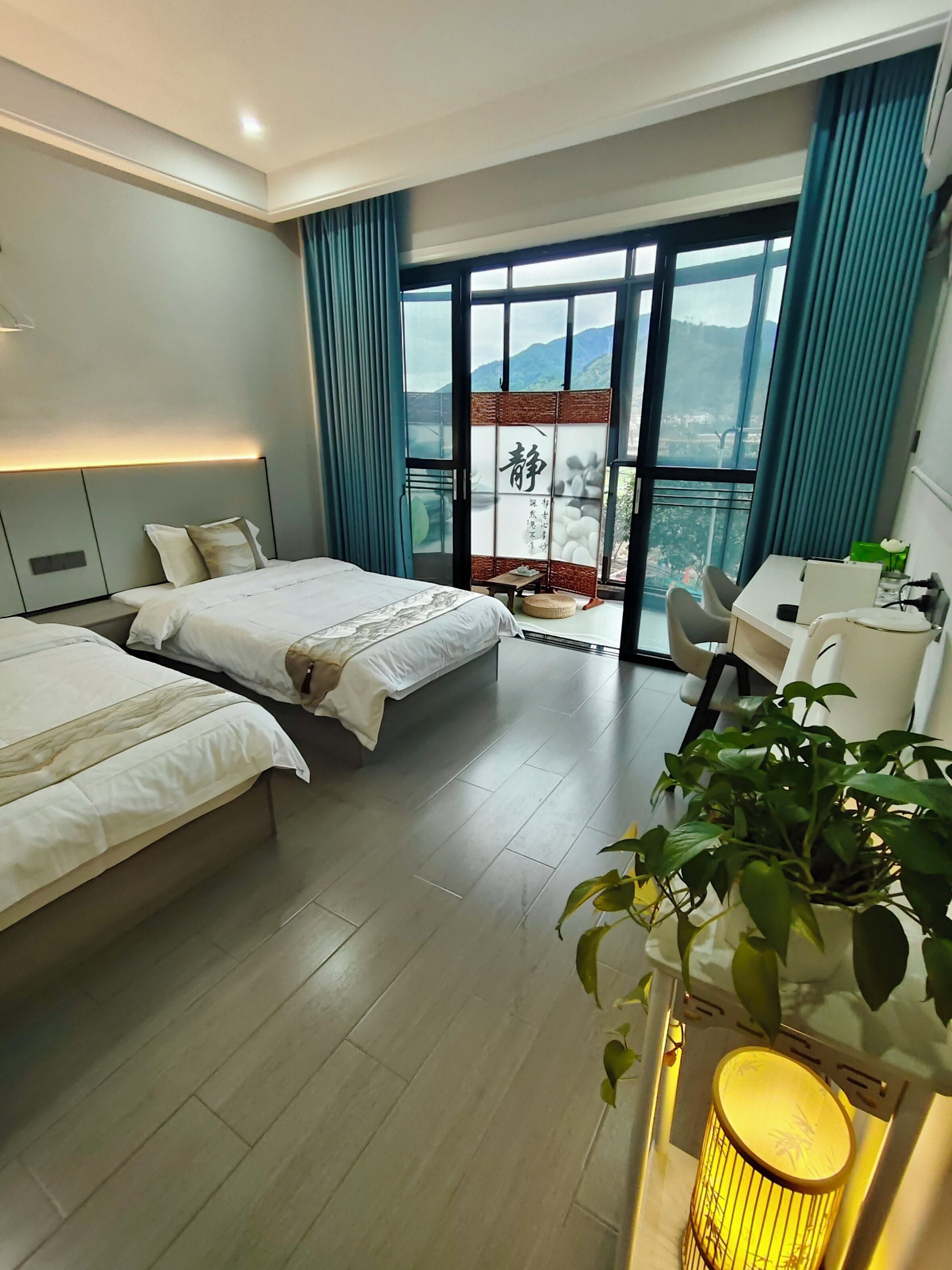 A modern hotel room with two beds, a desk, a chair, a potted plant, and a view of mountains through large windows.