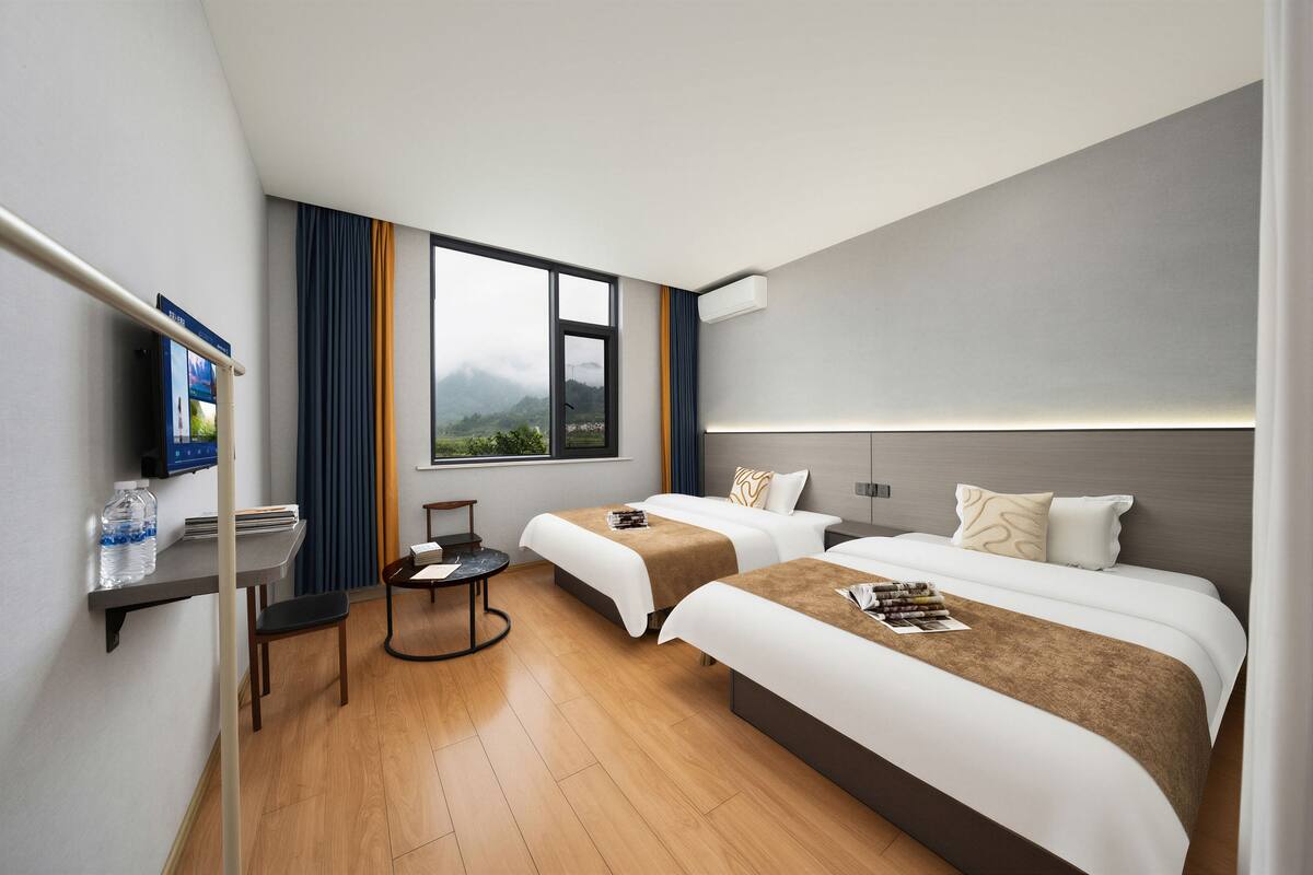 Habitación de hotel con dos camas, un escritorio, una silla, un televisor y ventana con vista a las montañas.