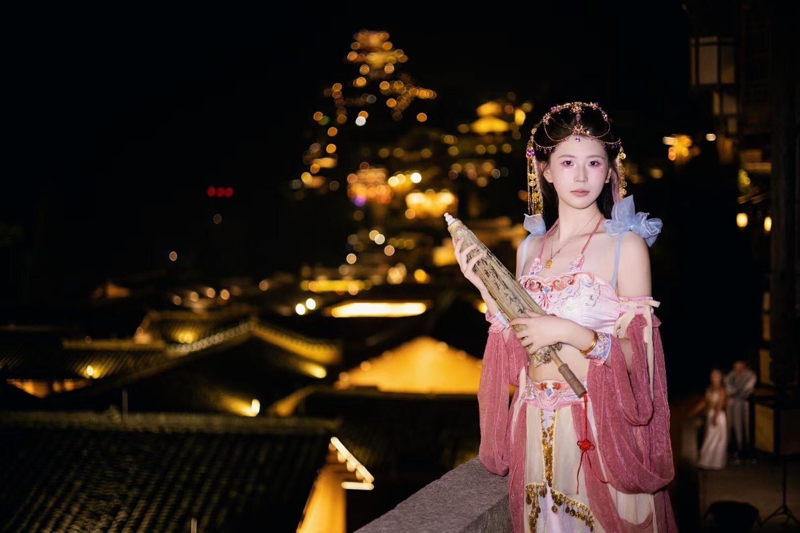 Una persona vestida con atuendo tradicional sostiene un instrumento musical, de pie en un balcón con vista a arquitectura tradicional iluminada de noche.