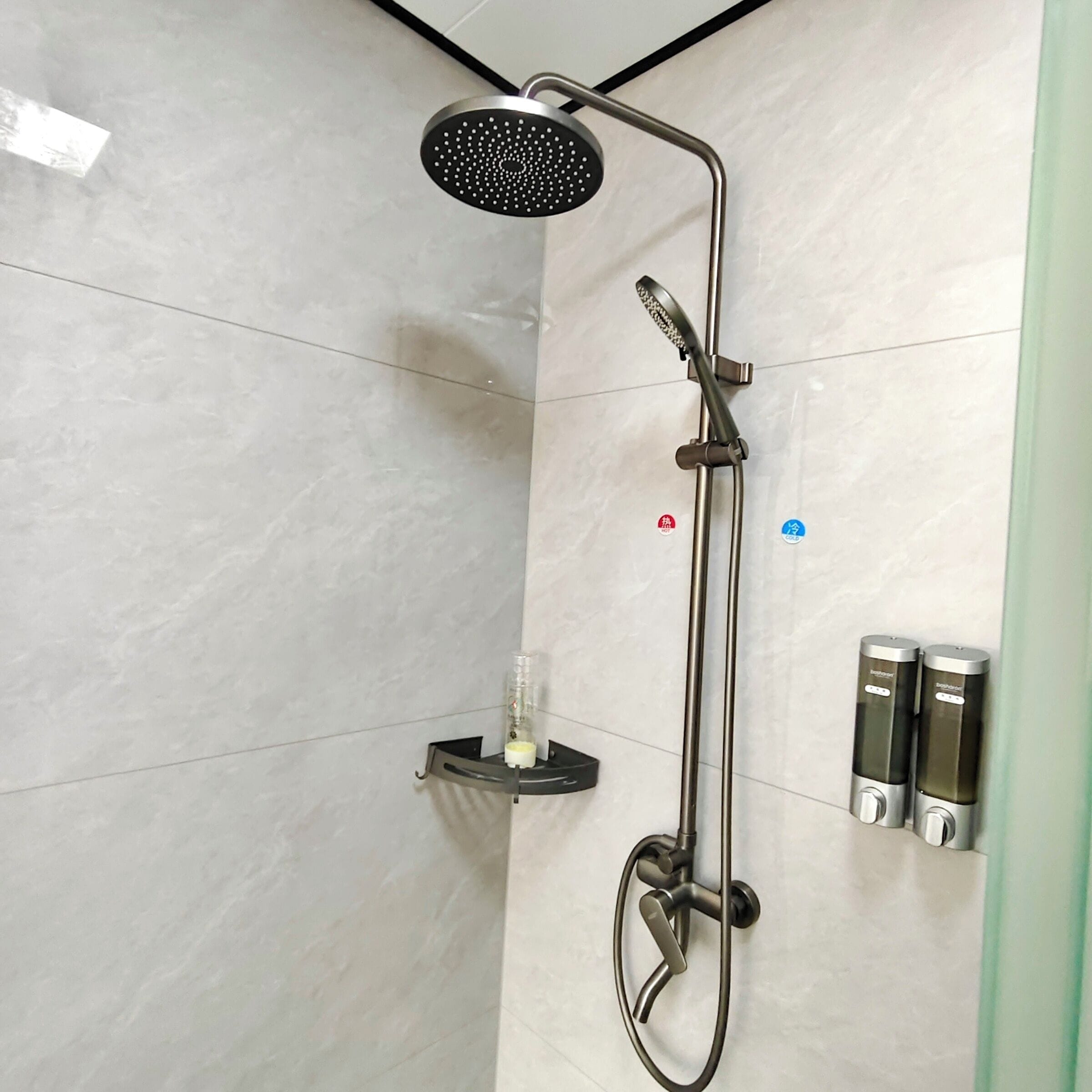 A modern shower with a handheld showerhead, a handheld scrubber, and two soap dispensers on a wall-mounted shelf.