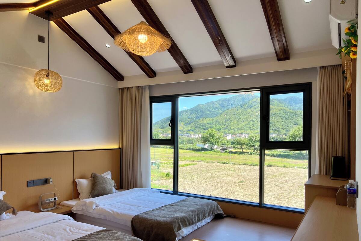 Una habitación de hotel con dos camas, un ventanal con vistas a la montaña, vigas de madera en el techo y una mesita de noche con lámpara.