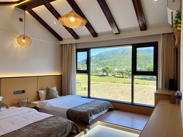 Une chambre d’hôtel avec deux lits, une grande fenêtre donnant sur les montagnes, des poutres en bois au plafond et une table de chevet avec une lampe.