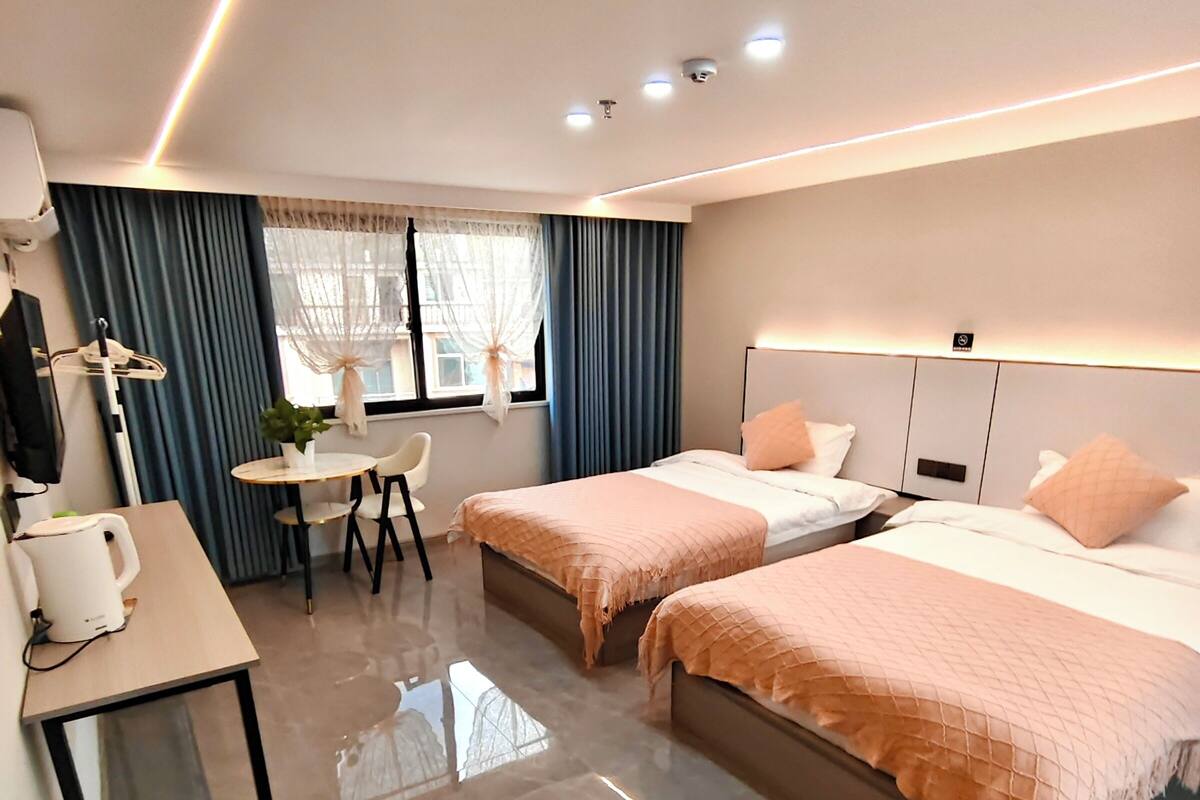 Habitación de hotel con dos camas, un escritorio con silla, una mesita y una ventana con cortinas.