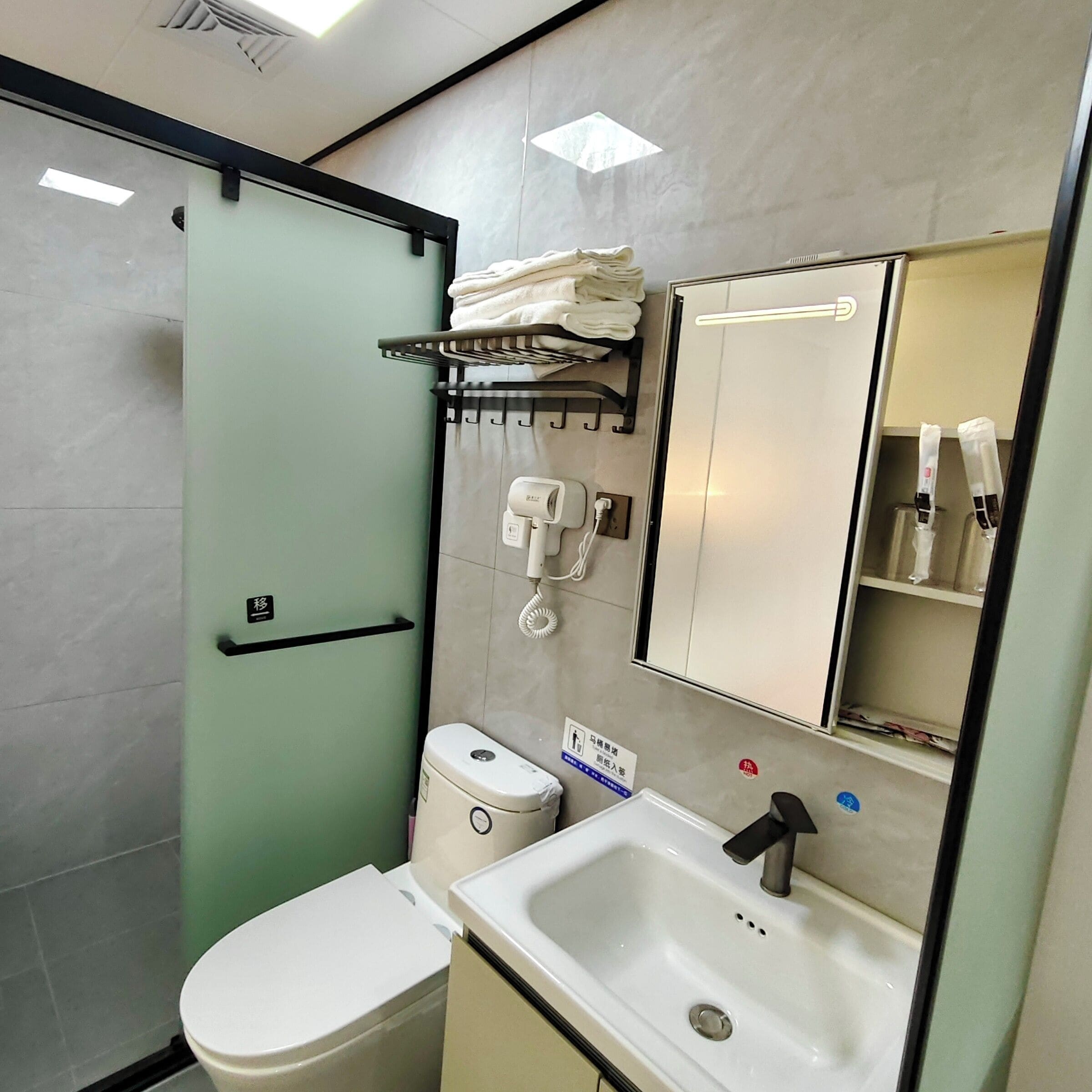 A bathroom with a toilet, sink, and a glass shower enclosure. There is a towel rack above the sink and a mirror above the toilet.